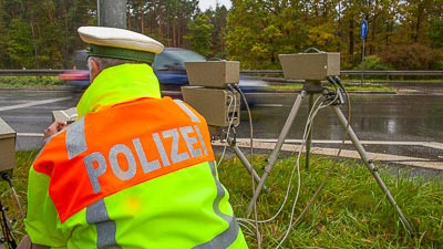 An einer Straße ist ein Gerät zur Geschwindigkeitskontrolle aufgebaut. Ein Polizist prüft das Gerät.