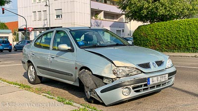  Straßenrand steht ein Unfallfahrzeug. Die vordere linke Seite ist kaputt, die Stoßstange hängt herunter und das Rad steht verdreht im Kotflügel.