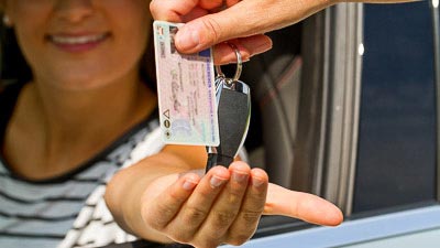 Eine junge Frau sitzt in einem Fahrzeug und hält die Hand auf. Eine andere Hand hält einen Führerschein und einen Autoschlüssel in der Hand und überreicht ihr die Gegenstände.