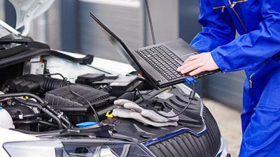 An einem Auto mit geöffneter Motorhaube steht ein Mechaniker mit Laptop. Dieser ist mit Kabel am Motor verbunden.