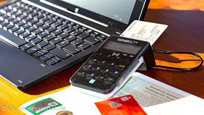 Laptop, Kartenleser mit Personalausweis, Kfz-Brief und EC-Karte auf einem Esstisch.