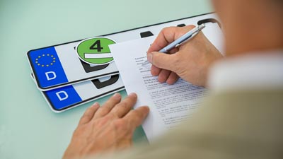 Kennzeichenschilder und eine Umweltplakette liegen auf dem Tisch. Eine Person füllt eine Vollmacht mit dem Stift aus.